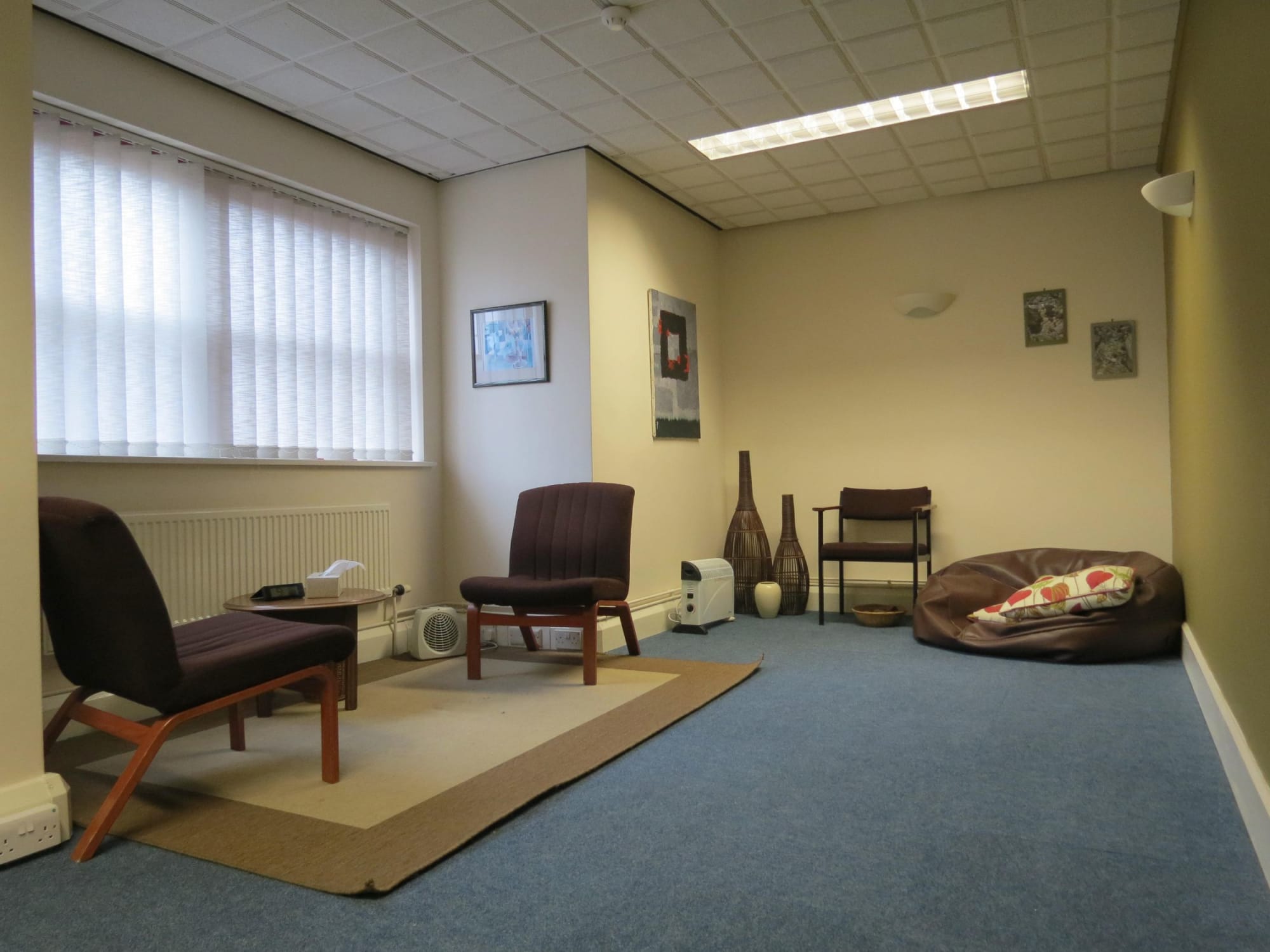 Image shows a large therapy room at Nottingham Counselling Service. There are two comfy chairs facing each other over a small coffee table in front of a window letting in natural light to the left. To the right there is another chair and a brown leat